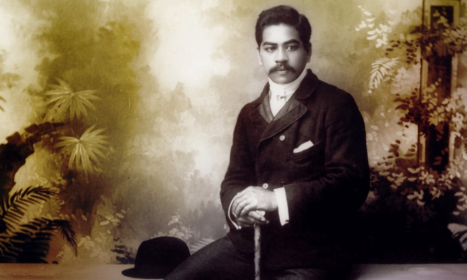 photo of a young Prince Kuhio, seated in front of a painted backdrop and holding a cane in formal Edwardian clothes with bowler