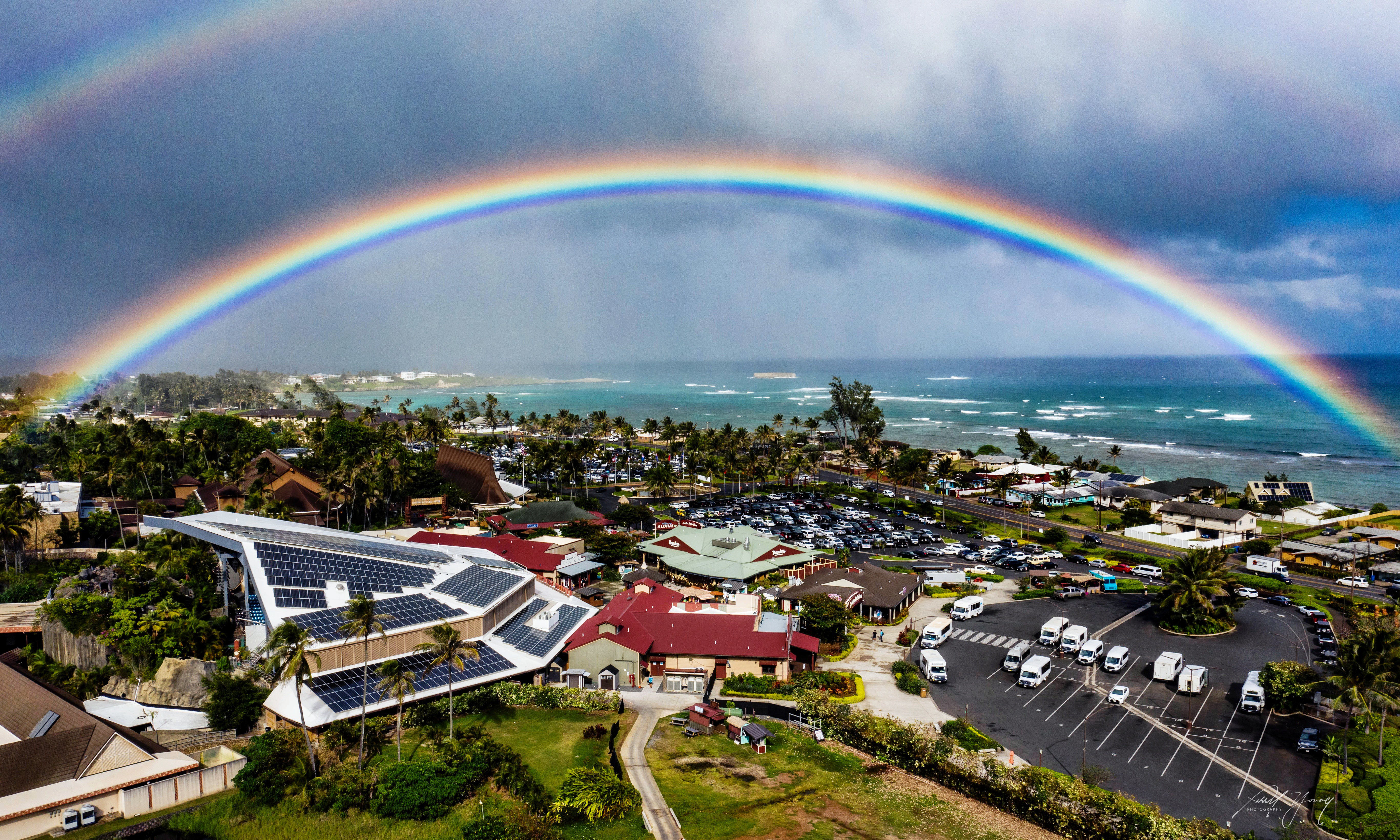 image shows the skyline surrounding the Polynesian Cultural Center with a viibrant rainbow and the ocean beyond