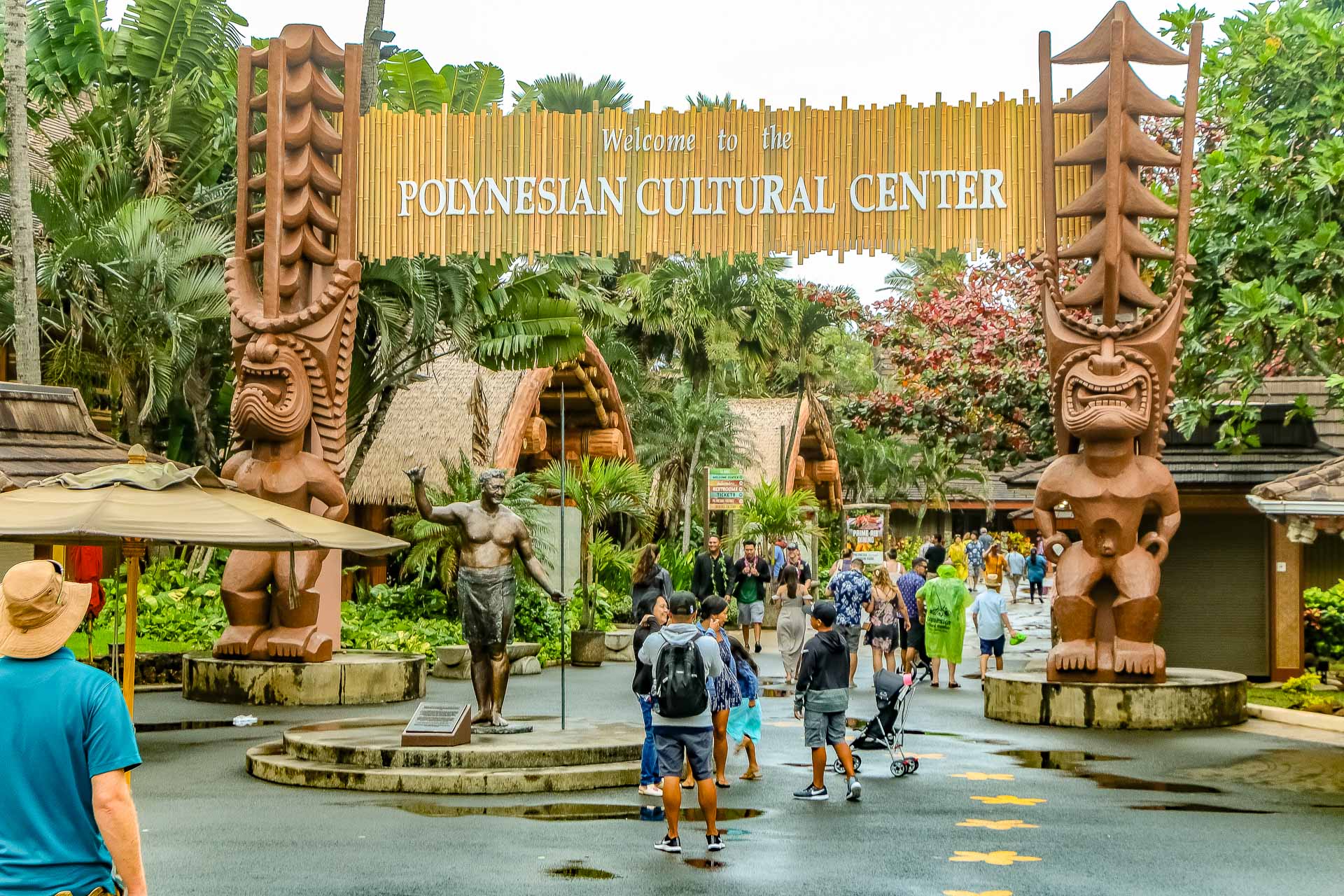 Visitors enter the Polynesian Cultural Center in Oahu, welcomed by towering tiki statues and lush tropical surroundings.