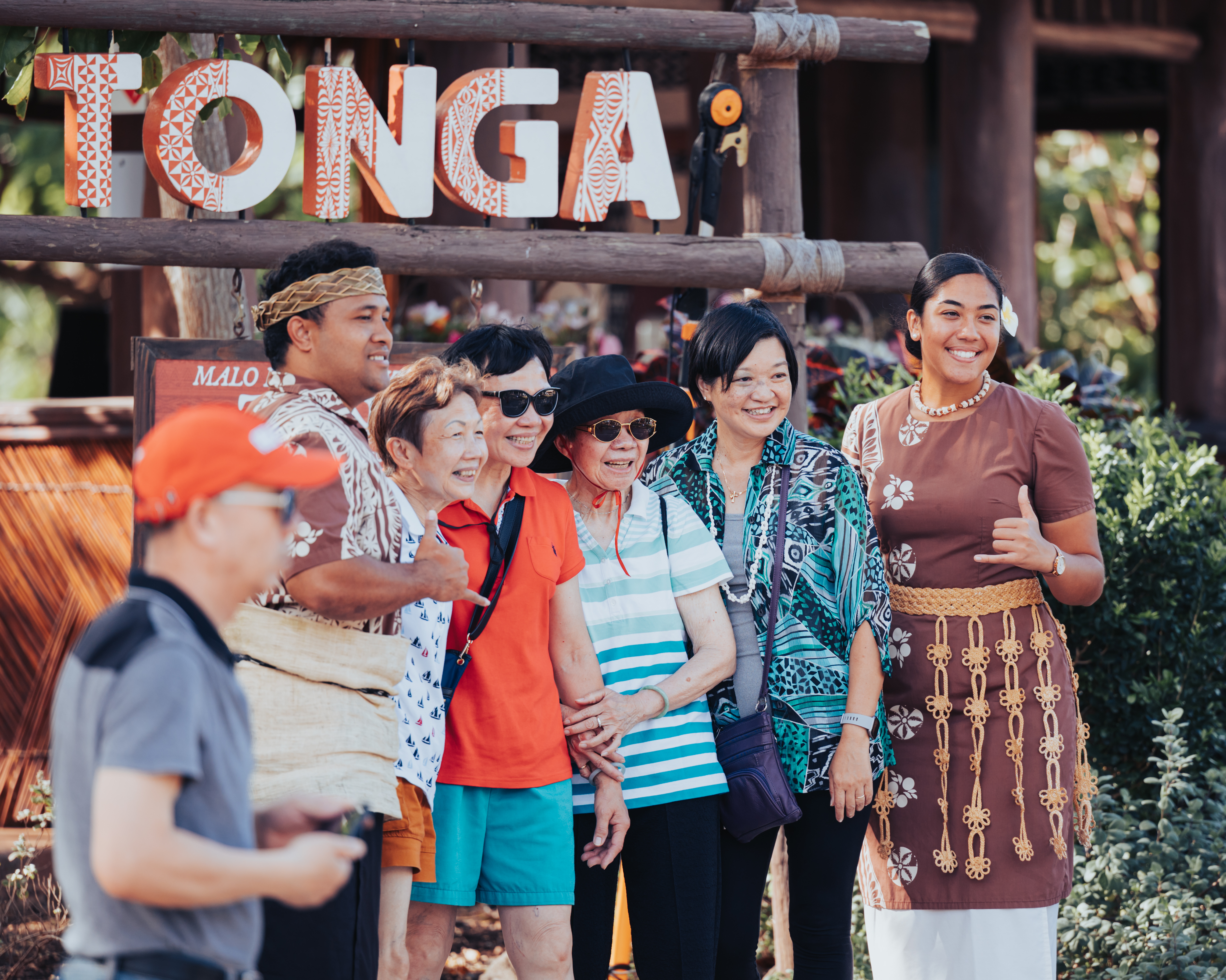 photo of guests taking a picture with Tongan village performers in front of Tonga village sign. A great example showcasing the love for our Ohana to all guests visiting the Polynesian Cultural Center.