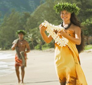 An image of a young woman and man showcases the Hawaiian word Aloha. Both wear Hawaiian attires, hakus (headpiece), kukui nuts necklaces, and traditional Hawaiian leis. 