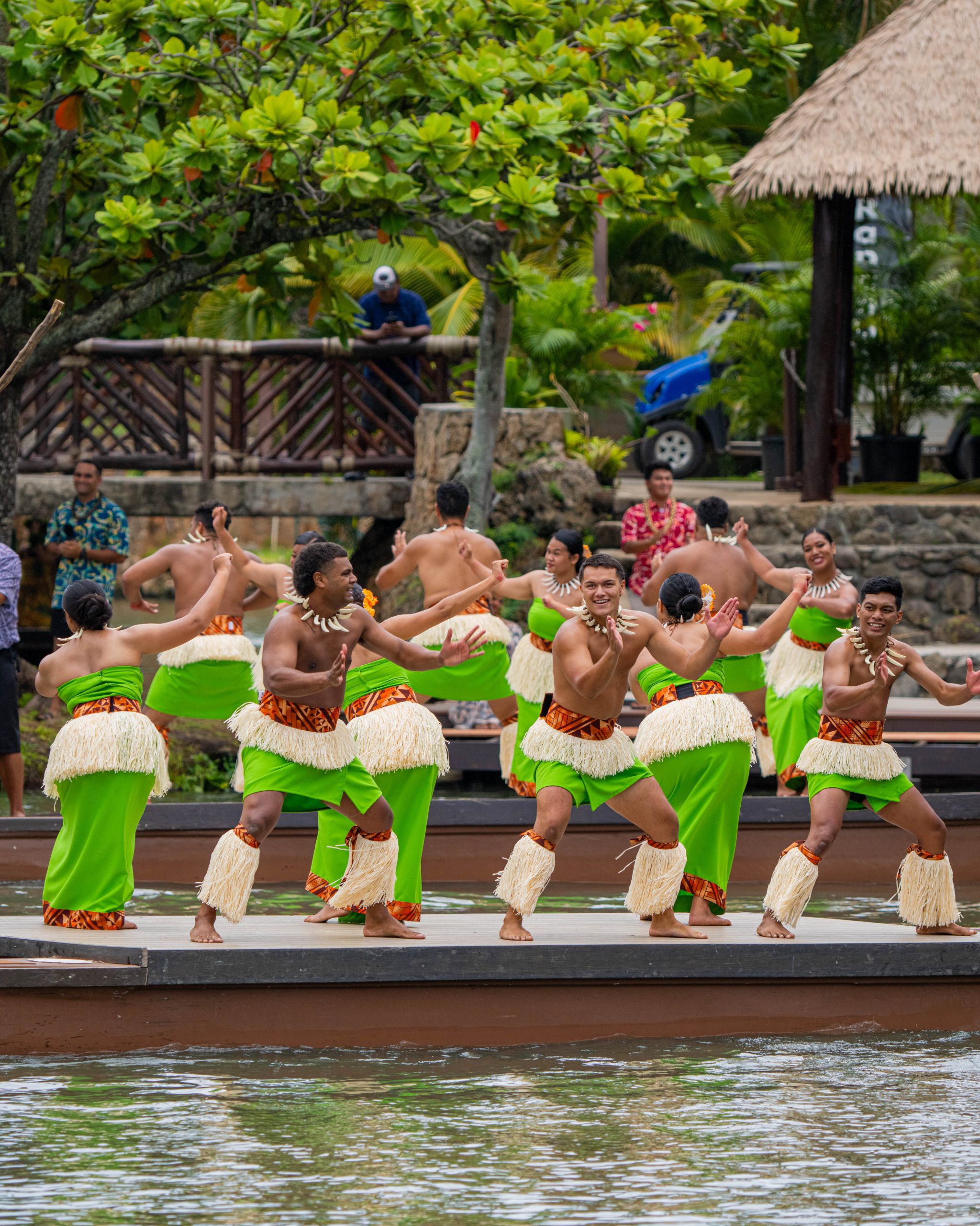 A group of men and women dancing on a canoe wearing bright green lavalava (wrap arounds) and puletasi (women’s attire).