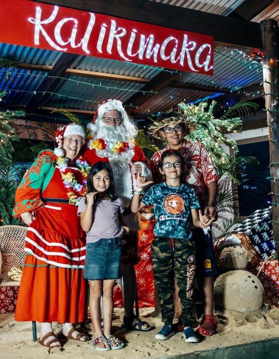 Red "Kalikimaka" banner hangs over Santa and Mrs. Claus, and a happy girl, boy, and man.