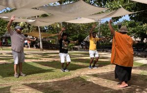 Under billowing canopies, a male worker in an orange, loose top and a sulu, teaches 3 male guests to dance.