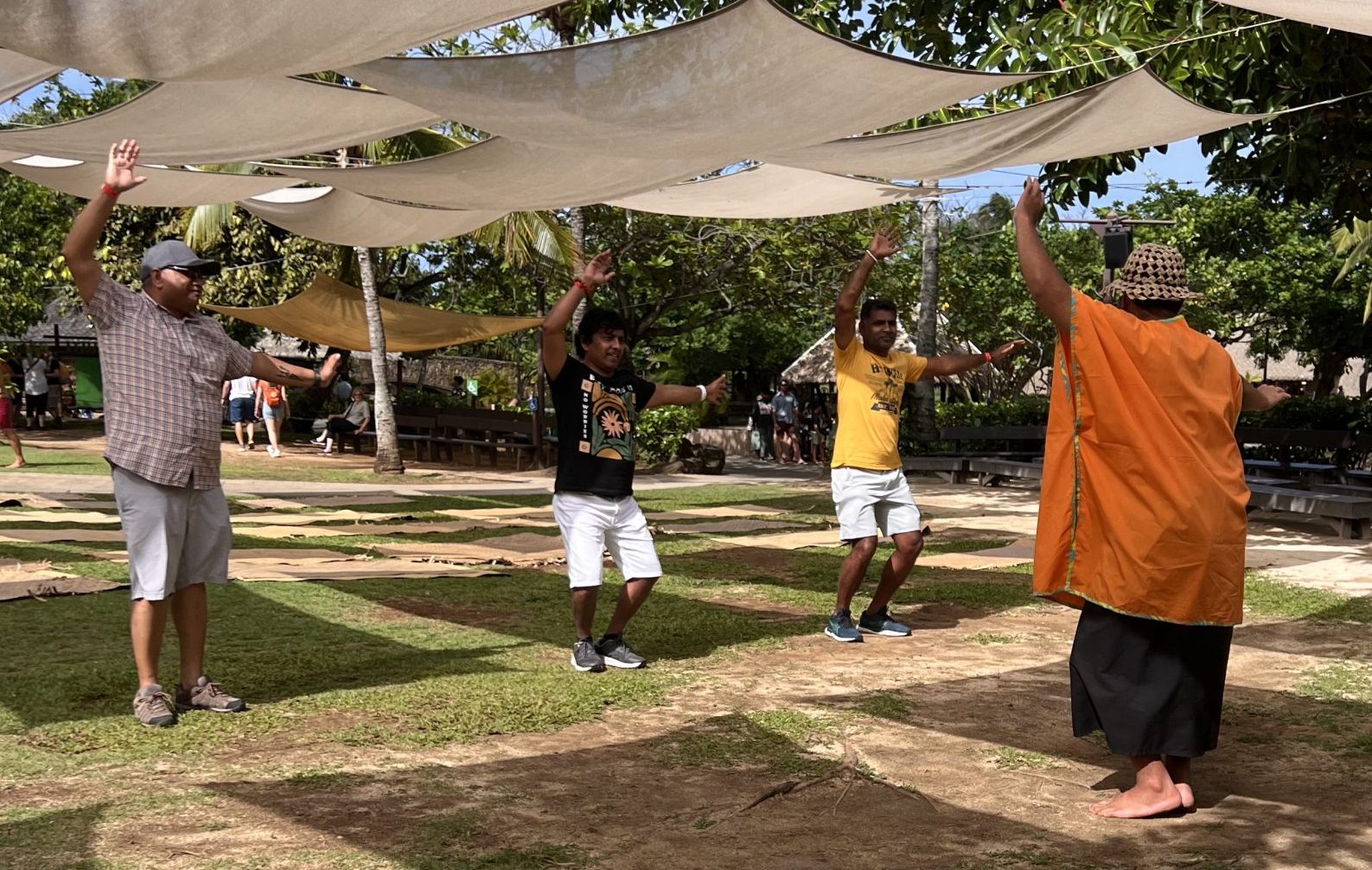 Under billowing canopies, a male worker in an orange, loose top and a sulu, teaches 3 male guests to dance.