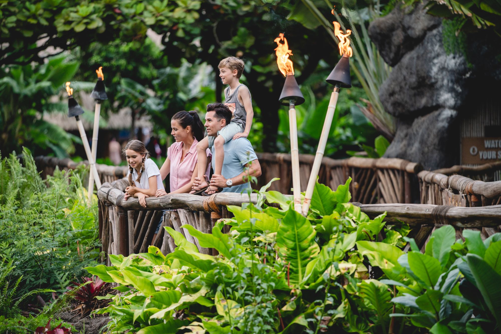 Parents and 2 kids on a rustic bridge, enjoying the lush vegetation. Boy sits on father's shoulders.