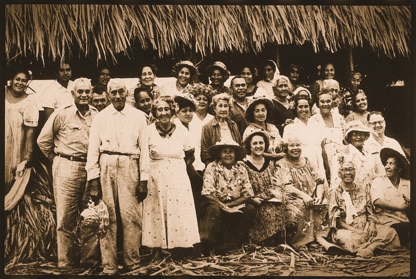 A vintage picture of people gathered and smiling in front of a large, thatched building.