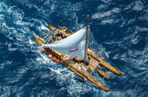 Double-hulled canoe viewed from overhead while it sails. The sail is filled with wind and crew members work on board.