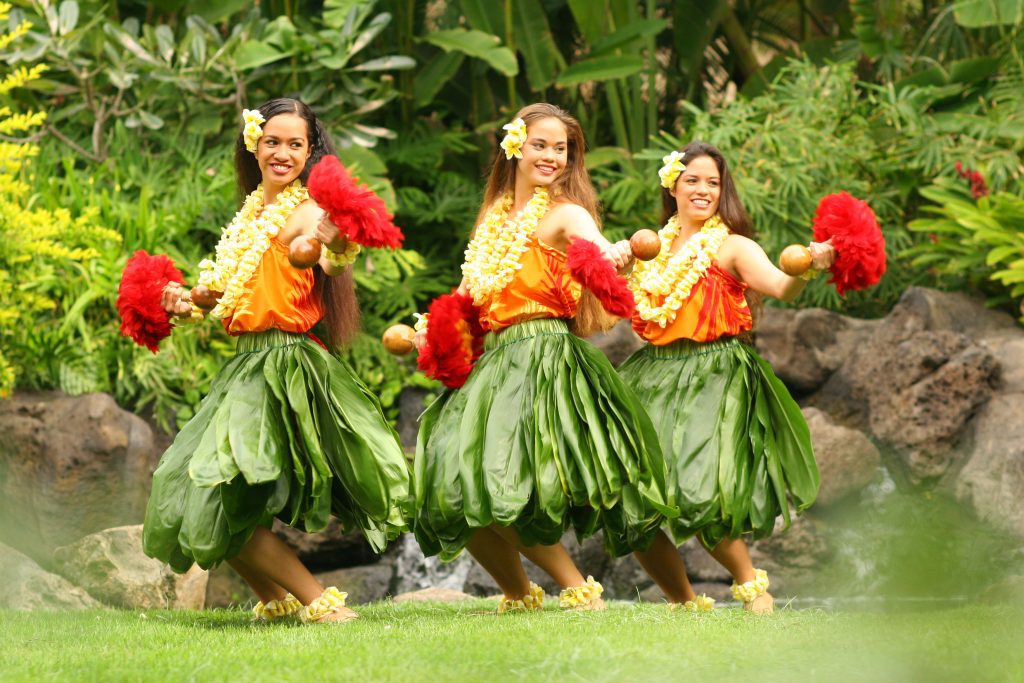 Three female hula dancers in ti leaf skirts, yellow tops and yellow lei.