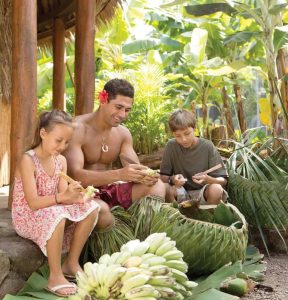 Male islander in costume helps 2 children peel bananas.