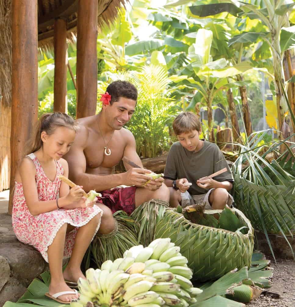 Male islander in costume helps 2 children peel bananas.